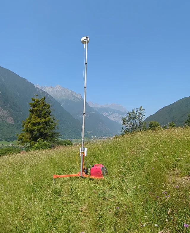 Richtstrahl-Station an einem abgelegenen Standort auf einer Wiese: ausgefahrener Mast mit Antenne auf einem orangen Dreibein-Stativ, daneben ein rotes Stromaggregat, im Hintergrund Berge